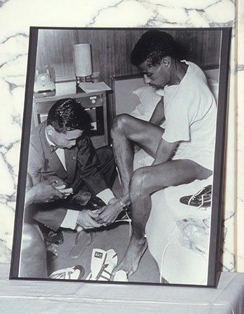 ホテルの部屋でアベベの足にシューズを合わせる鬼塚喜八郎（1992年にアシックスがアベベの妻と娘に寄贈した写真）